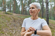 © Anatoliy Karlyuk - Close up view of self determined middle aged sportswoman in white t-shirt adjusting smart watch, checking fitness statistics, monitoring her running performance during cardio workout in park