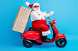 © deagreez - Full length profile side photo of crazy white grey hair bearded santa claus drive x-mas christmas scooter deliver pizza stack point finger way wear red costume isolated blue color background