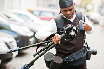 Wall Mural - Young professional african american videographer holding professional camera with tripod pro equipment. Afro cameraman wearing black duraq making a videos.