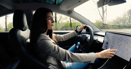 New York - October 10, 2020: Beautiful Caucasian woman sitting in electric vehicle Tesla Model 3 and typing on on-board touch screen. Female driver traveling in car and tapping on dashboard computer