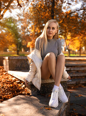 Beautiful student girl with perfect legs sitting on the parapet in the autumn park in white sneakers and coat