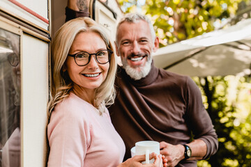 Close up shot of pleased senior caucasian couple smiling at the camera with cups of coffee standing in the van doorway