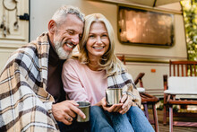 Pleased European Mature Couple Sitting Together On The Van Porch Covered With Blanket With Mugs Of Coffee