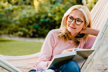 Pretty Aged Blonde Woman In Glasses Reading A Book In Hammock In Summer Garden