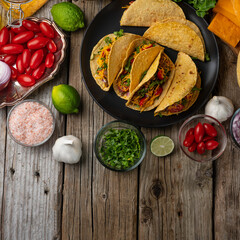 Wall Mural - Top view of tasty mexican tacos with meat on black round plate on rustic wooden table. Traditional mexican cuisine. Healthy ingredients for cooking meal background. Concept of street food.