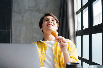 Asian man in yellow shirt working and eating snack in modern office.