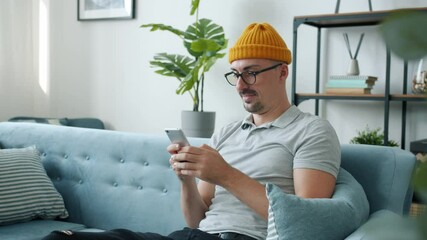 Slow motion of happy guy using smartphone texting laughing enjoying communication sitting on couch at home. People and modern devices concept.
