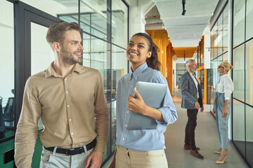 Wall Mural - Two young happy multiracial male and female coworkers talking and smiling while walking in the modern office