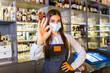 © Graphicroyalty - Happy young woman wearing protective face mask in the retaurant during coronavirus pandemic, Bartender showing Ok sign. Shelves full of bottles with alcohol on the background