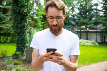 Young bearded man standing outdoor with a mobile phone in his hand. Receiving a message on a cell phone, smartphone.