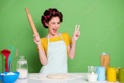 Portrait of her she nice attractive pretty cheerful cheery funky housewife cooking delicious cake pizza rolling dough winking showing v-sign isolated over green pastel color background