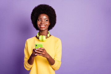 Sticker - Photo of positive dark skin lady hold cellphone look empty space wear yellow jumper headphones isolated on purple color background