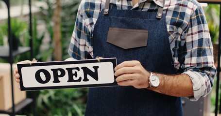 Wall Mural - Close up of young Caucasian male owner of florist's shop holding Open sign in hands and smiling at camera. Happy handsome man flower center worker in apron and mask standing indoors. Business concept