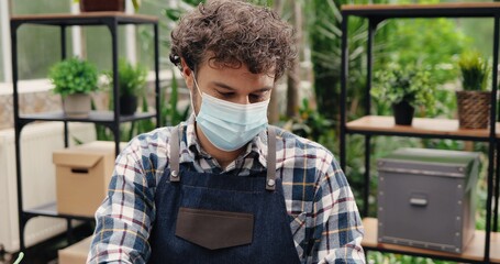 Wall Mural - Close up of young male entrepreneur in flower shop sitting at table and typing on laptop. Portrait of handsome Caucasian joyful man greenhouse worker in mask browsing on computer. Job concept