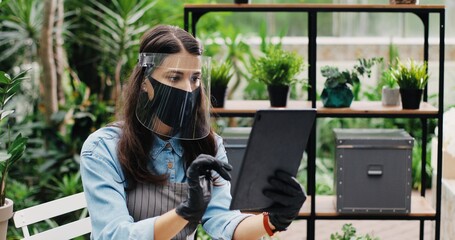 Wall Mural - Portrait of Caucasian female entrepreneur sitting at table flower shop and tapping and typing on tablet indoor. Beautiful woman owner of greenhouse in face shield browsing on device. Florist concept