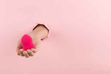 Wall Mural - Girl hand holds a beauty blender for makeup from a hole in a pink background, copy space.