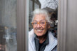 older woman wearing eyeglasses smiling through window behind closed door during covid19 quarantine