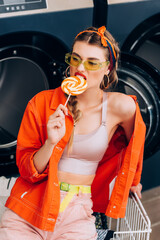 Sticker - trendy young woman in sunglasses biting lollipop near cart and washing machines in laundromat