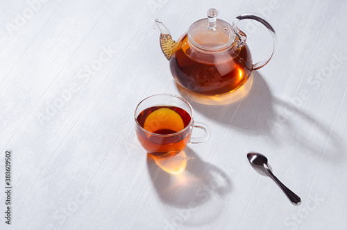 Sunny tradition teatime with black tea in transparent teapot with cup in bright cozy orange sun beams with shadows white wood table top view, copy space.