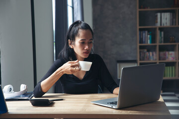 Wall Mural - Remote working at Home office Concept. Businesswoman working online via laptop and drink coffee. Asian young entrepreneur watching webinars and talking during meeting video conferences calls with team