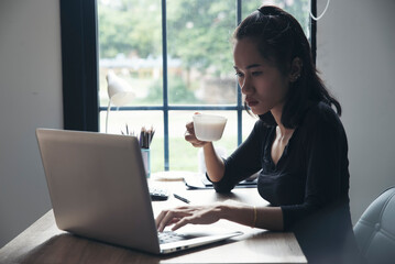 Wall Mural - Remote working at Home office Concept. Businesswoman working online via laptop and drink coffee. Asian young entrepreneur watching webinars and talking during meeting video conferences calls with team