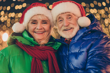 Wall Mural - Two retired pensioner grey white hair people affectionate couple woman man celebrate x-mas christmas jolly event walk outside under illumination wear santa claus headwear