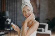 Leinwandbild Motiv Indoor shot of young smiling woman applies moisturizer cream on face, takes care of her skin and complexion, puts lotions, has minimal makeup, wrapped in bath towel. Beauty, cosmetology concept