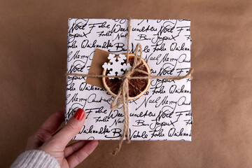 A woman's hand is holding the gift on a brown background, the box is decorated with elements of Christmas decor, brown twine, snowflake, card for the inscription