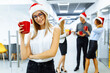 © Shopping King Louie - Happy young business woman in Santa Claus hat, holding a red mug, with a group of colleagues in the background, during a coffee break in the office. Meeting New Year and Christmas