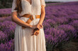 A girl in a long dress holds a sprig of lavender. There's a lavender field in the background. A cropped image.