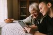 © Dragica - A cute gray-haired retired grandmother and her teenage caucasian granddaughter look at a tablet as they sit at a table in their grandmother's house. Grandma's education about new technology
