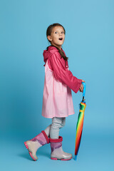 Wall Mural - emotional surprised cute child girl with multicolored umbrella in pink rain coat and rubber boots on blue background.