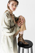 © LIGHTFIELD STUDIOS - trendy young woman in trench coat and scarf posing near chair on white