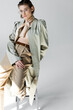 © LIGHTFIELD STUDIOS - trendy young woman in trench coat and scarf sitting on stool while posing on grey
