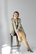 © LIGHTFIELD STUDIOS - full length of young woman in trench coat and scarf sitting on stool while posing on grey