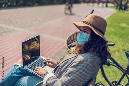 Woman in a hat and medical mask works sitting on a park bench. New normal concept.