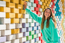 Smiling Young Woman Standing By Colorful Brick Wall On Sunny Day