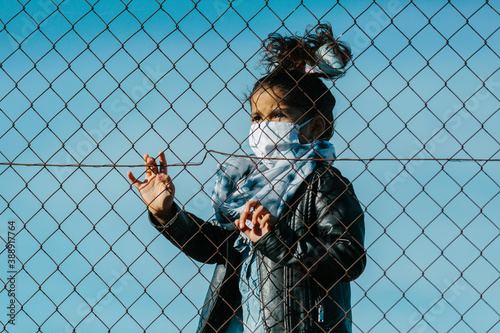 Fotografía Latin young girl wearing a mask, looking away with an serious expression, behind a fence, in a blue sky background
