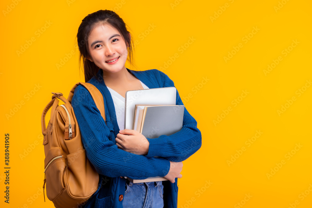 Happy asian female college student smiling at camera on yellow background and copy space, holding laptop and books, hanging bagpack. Youth girl student is exchange student. education concept