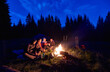 © anatoliy_gleb - Evening camping near fire, spruce forest on background. Group of four friends sitting together near campfire, enjoying fresh air near tent with tourists equipment nearby, dark relaxing atmosphere.