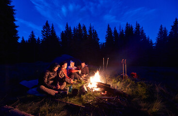Wall Mural - evening camping near fire, spruce forest on background. group of four friends sitting together near 