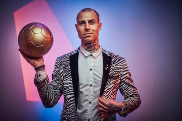 Positive and succesful football trainer dressed in custom suit with jewellery poses with golden ball in abstract light background.