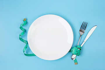 Wall Mural - Top view Green Measuring tape wrapped around fork and knife with white ceramic dish on blue background. dieting, weight loss, obesity and food control concept