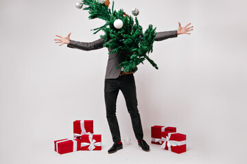 Man in black pants having fun during studio new year photoshoot. Indoor portrait of carefree guy posing with green christmas tree decorated with balls.