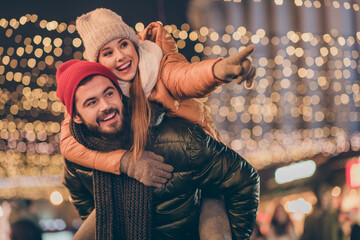 Wall Mural - Photo of two friends students guy hug piggyback girl point finger under evening x-mas illumination outdoors