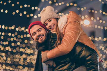 Poster - Photo of jolly guy hugging piggyback his beloved girlfriend under x-mas evening outdoors illumination wear season coats