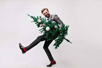 Laughing young man in trendy costume enjoying new year party. Smiling office worker holding christmas tree.