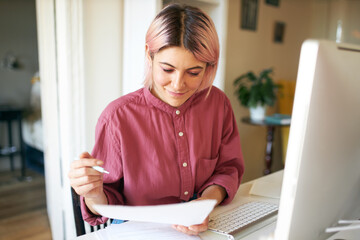 Creative confident young woman sitting in front of computer monitor with papers in her hands, filling in application form, searching for new job. Pretty student girl doing research for course paper