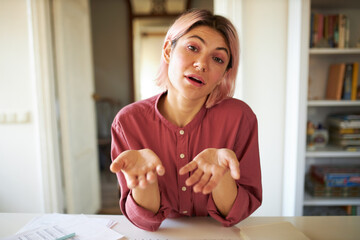 Beautiful stylish young female with bob pinkish hair sitting at table reaching out hands and opening mouth, conducting individual online lesson tutorial, teaching foreign language using web camera