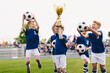 © matimix - Happy boys in elementary school sports team celebrating soccer succes in tournament final game. Kids winning football game. Happy children sports team rising golden trophy